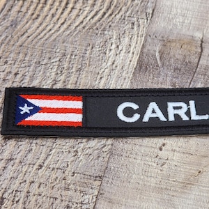 May include: A black patch with the name "CARLOS" in white block letters. The patch includes the flag of Puerto Rico on the left side, featuring a white star, red stripes, and blue triangle.