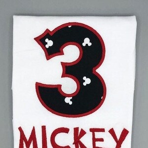 Puede incluir: Camiseta blanca con un número 3 negro con contorno rojo y cabezas de Mickey Mouse blancas. El texto "MICKEY" está en rojo debajo del número.