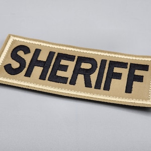 May include: A tan fabric patch with a white border and the word "SHERIFF" embroidered in black.
