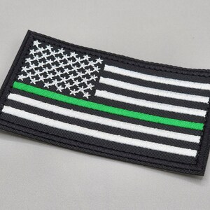 May include: Black and white American flag patch with a green stripe across the middle. The patch is embroidered and has a black border.