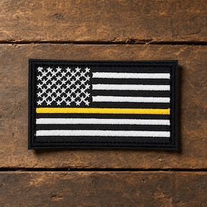 Könnte beinhalten: Ein rechteckiger bestickter Aufnäher mit einem stilisierten amerikanischen Flaggendesign in Schwarz und Weiß. Die Flagge zeigt ein Feld mit weißen Sternen auf schwarzem Grund und horizontale Streifen sowie einen gelben Streifen.