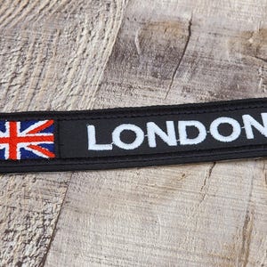 Op de afbeelding: Zwart rechthoekig embleem met het woord "LONDON" in witte letters. De Union Jack vlag staat links. Geschikt om op kleding of tassen te naaien.