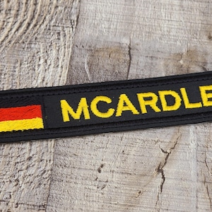 May include: A black embroidered patch featuring the name "MCARDLE" in yellow, with the German flag on the left. The rectangular patch has a black border and is designed for application.
