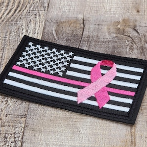 May include: Embroidered patch featuring a stylized American flag with a pink line and a pink ribbon. The flag has a black border, white stars, and white and black stripes. The pink ribbon is a symbol of breast cancer awareness.