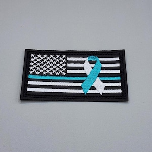 May include: A rectangular embroidered patch featuring a stylized American flag design with black and white horizontal stripes and a field of white stars. A single horizontal teal stripe runs across the center, overlaid with a teal and white awareness ribbon.