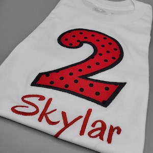 Puede incluir: Camiseta blanca con un número dos rojo con lunares negros y el nombre "Skylar" bordado en rojo.