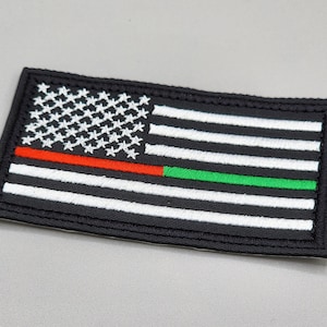 Puede incluir: Un parche bordado negro con un diseño de bandera estadounidense blanca. La bandera tiene una franja roja y una franja verde añadidas al diseño.