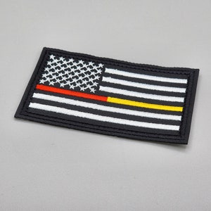 Puede incluir: Un parche bordado negro con una franja roja y amarilla a través de la bandera estadounidense. La bandera es blanca con rayas rojas y blancas y 50 estrellas blancas sobre un campo azul.