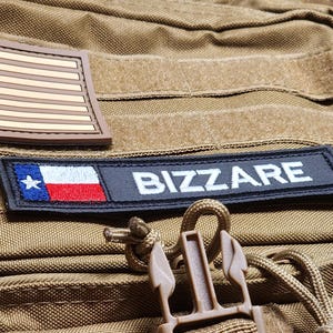Puede incluir: Primer plano de una mochila táctica color canela con dos parches. Un parche muestra la bandera estadounidense, el otro dice "BIZZARE" sobre la bandera del estado de Texas. La mochila tiene una hebilla y detalles de cuerda.