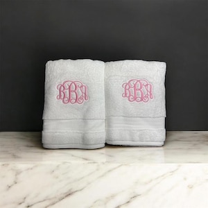 May include: Two white bath towels with a pink embroidered monogram. The towels are folded and stacked, with a decorative band near the bottom. The background is a dark gray, and the towels are on a marble surface.