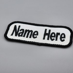 May include: White fabric patch with black embroidered text that reads "Name Here".