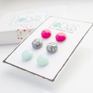 May include: A set of six stud earrings on a white card with the text "Lobos handmade studs". The earrings are in three different styles: two pink, two silver glitter, and two light blue.