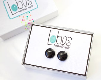 Puede incluir: Un par de pendientes de botón negros en una pequeña caja blanca con el texto "Lobes handmade studs" en la caja.