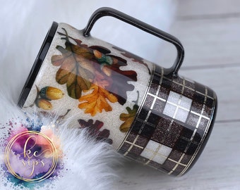 May include: A white ceramic mug with a black handle. The mug is decorated with a fall-themed design of leaves and acorns in shades of brown, orange, and yellow. The bottom half of the mug is decorated with a black and white plaid pattern.