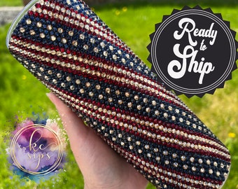 May include: A clear tumbler with a red, white, and blue American flag design covered in rhinestones. The tumbler is held in a hand. The text "Ready to Ship" is in a circle in the background.