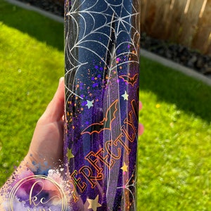 K&ouml;nnte beinhalten: Ein schwarzer und lilafarbener Glitzerbecher mit einem Spinnennetz-Design, Flederm&auml;usen und goldenen Sternen. Der Becher hat eine klare, glitzernde Oberfl&auml;che und der Text "Perfectly" ist in Orange an der Seite geschrieben.