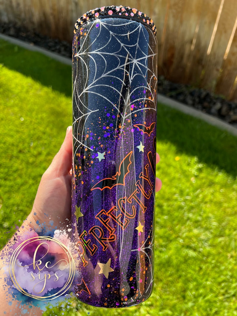 K&ouml;nnte beinhalten: Ein schwarzer und lilafarbener Glitzerbecher mit einem Spinnennetz-Design, Flederm&auml;usen und goldenen Sternen. Der Becher hat eine klare, glitzernde Oberfl&auml;che und der Text "Perfectly" ist in Orange an der Seite geschrieben.