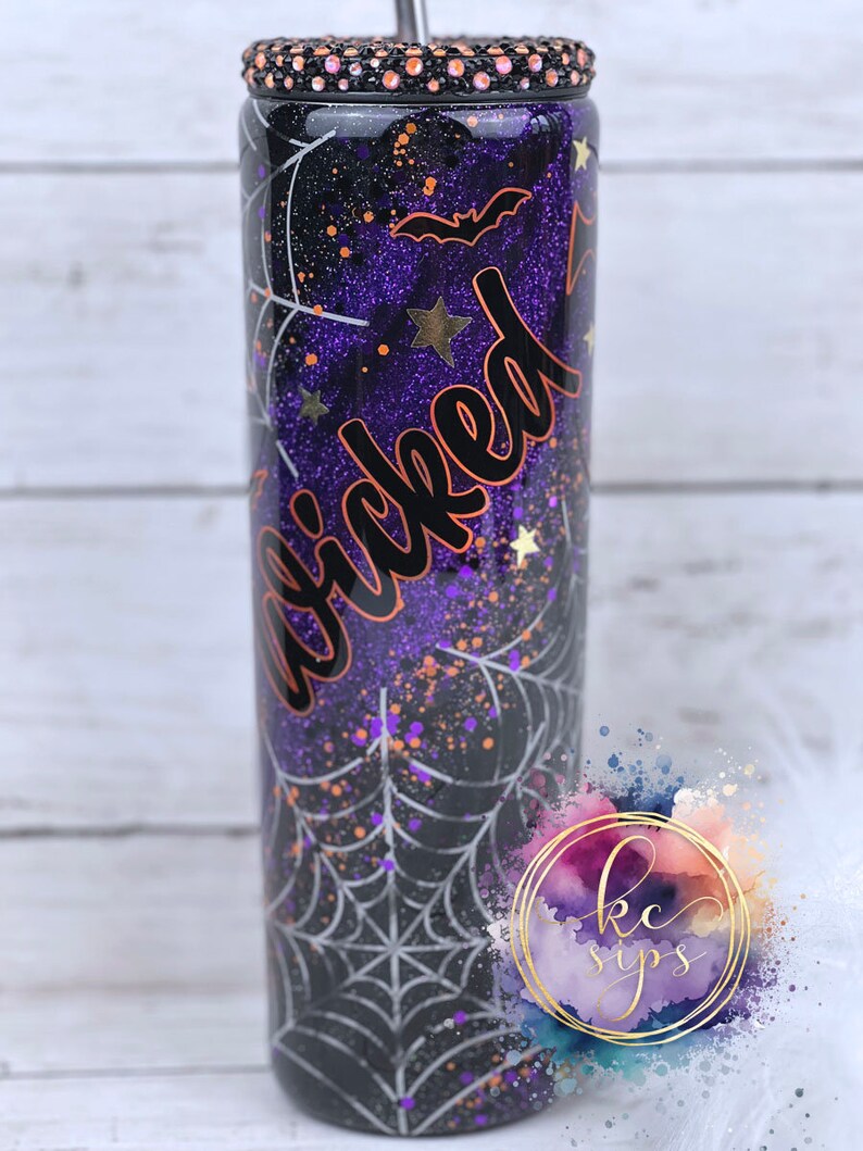 K&ouml;nnte beinhalten: Ein schwarzer und lilafarbener Becher mit einem Spinnennetz-Design und dem Wort "Wicked" in orangefarbener Schrift. Der Becher hat eine glitzernde Oberfl&auml;che und einen schwarzen Deckel mit Strasssteinen.
