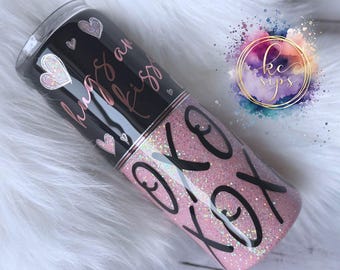 May include: A black and pink tumbler with glitter. The tumbler has the words "hugs and kisses" written on it in a cursive font. The bottom of the tumbler has the words "XOXO" written on it in a black font.