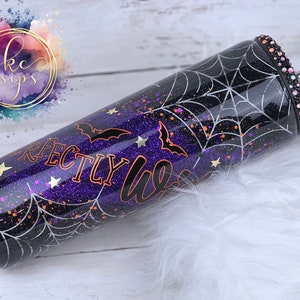 K&ouml;nnte beinhalten: Ein schwarzer und lilafarbener Glitzerbecher mit einem Spinnennetz-Design und dem Text "Perfectly Lit" in kupferfarbenem Glitzer. Der Becher hat einen durchsichtigen Deckel mit einem silbernen Strohhalm und einem Strassstein-Akzent.