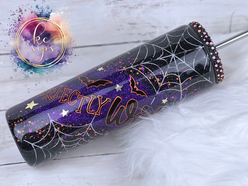 K&ouml;nnte beinhalten: Ein schwarzer und lilafarbener Glitzerbecher mit einem Spinnennetz-Design und dem Text "Perfectly Lit" in kupferfarbenem Glitzer. Der Becher hat einen durchsichtigen Deckel mit einem silbernen Strohhalm und einem Strassstein-Akzent.