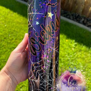 K&ouml;nnte beinhalten: Ein schwarzer und lilafarbener Glitzerbecher mit einem Spinnennetz-Design und dem Wort "Wicked" in orangefarbenem Glitzer. Der Becher hat einen schwarzen Deckel mit Strasssteinen.