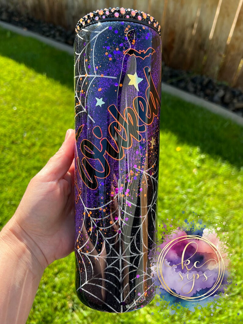 K&ouml;nnte beinhalten: Ein schwarzer und lilafarbener Glitzerbecher mit einem Spinnennetz-Design und dem Wort "Wicked" in orangefarbenem Glitzer. Der Becher hat einen schwarzen Deckel mit Strasssteinen.