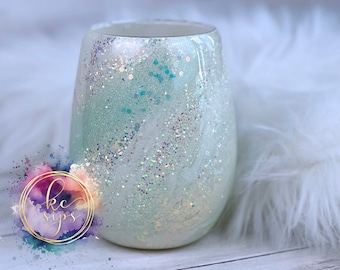 May include: A white tumbler with a light blue and iridescent glitter design. The tumbler is sitting on a white fluffy surface.