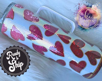 May include: White tumbler with a gold rim and a pink glitter heart pattern. The tumbler has a handle and is ready to ship.