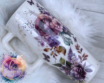 May include: White stainless steel tumbler with a floral design featuring purple, pink, and green flowers and leaves. The tumbler has a white handle and a glittery finish.