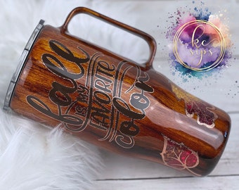 May include: A brown and gold stainless steel travel mug with a wood grain design. The mug has a handle and a lid. The text "Fall is my favorite color" is printed on the mug in a gold script font. The mug is decorated with gold glitter leaves.