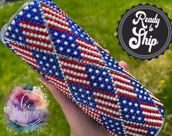 May include: A white tumbler with a red, white, and blue American flag design made of rhinestones. The tumbler is ready to ship.