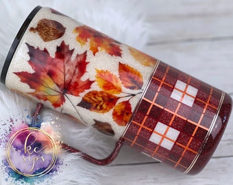 May include: A glittery tumbler featuring autumn leaves and an acorn design on a beige background. The bottom half has a red plaid pattern with copper accents. The tumbler has a black rim and a matching handle. The logo "kc sips" is visible.