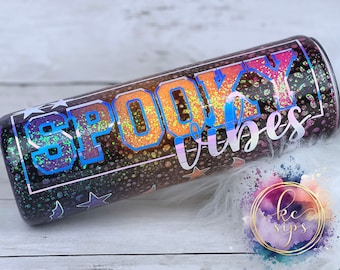 May include: A black and brown glitter tumbler with the text "Spooky Vibes" in rainbow iridescent letters. The tumbler has a black background with white stars and bats.
