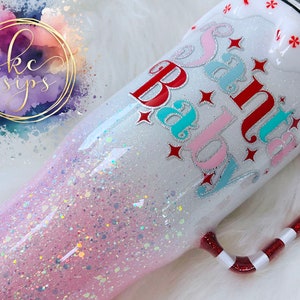 Könnte beinhalten: Ein weißer Becher mit einem rosa-weißen Glitzer-Ombre-Design. Der Becher hat einen roten und weißen Zuckerstangen-Griff und trägt die Aufschrift "Santa Baby" in roten, rosa und blauen Buchstaben.
