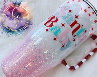 May include: White tumbler with a pink and white glitter ombre design. The tumbler has a red and white candy cane handle and says "Santa Baby" in red, pink, and blue lettering.
