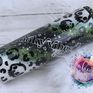 Könnte beinhalten: Ein schwarzer und weißer Becher mit einem grünen Glitzerdesign und den Worten "Spooky Vibes" in silberner Schrift. Der Becher hat ein schwarz-weißes Leopardenmuster und ein schwarz-weißes Spinnennetzdesign.
