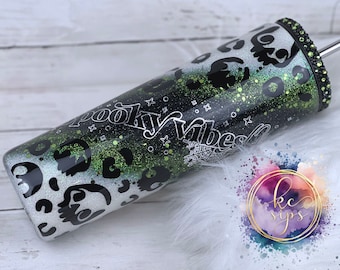 以下が含まれることがあります： 黒と白のタンブラーに緑のグリッターのデザインと銀色の文字で「Spooky Vibes」と書かれています。タンブラーには黒と白のレオパード柄と黒と白の蜘蛛の巣のデザインが施されています。