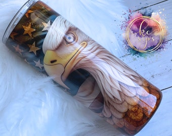 May include: A stainless steel tumbler with a patriotic design featuring a bald eagle with a close-up of its head and beak. The background is a faded American flag with stars and stripes.