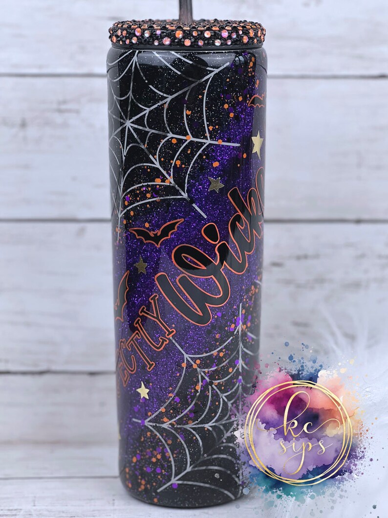 K&ouml;nnte beinhalten: Ein schwarzer und lila glitzernder Becher mit einem Spinnennetz-Design und dem Text "Perfectly Wicked" in Orange. Der Becher hat einen schwarzen Deckel mit Strasssteinen.