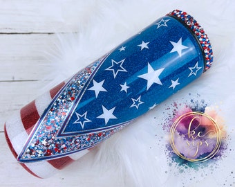 May include: A stainless steel tumbler with a red, white, and blue glitter design resembling the American flag. The tumbler has a clear lid with a bedazzled rim.