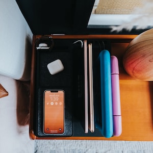 May include: A wooden bedside table with a black organiser tray holding a white wireless charging pad, a white case for wireless earbuds, a smartphone with a digital clock displaying 1:39, and a blue and purple case for a tablet.