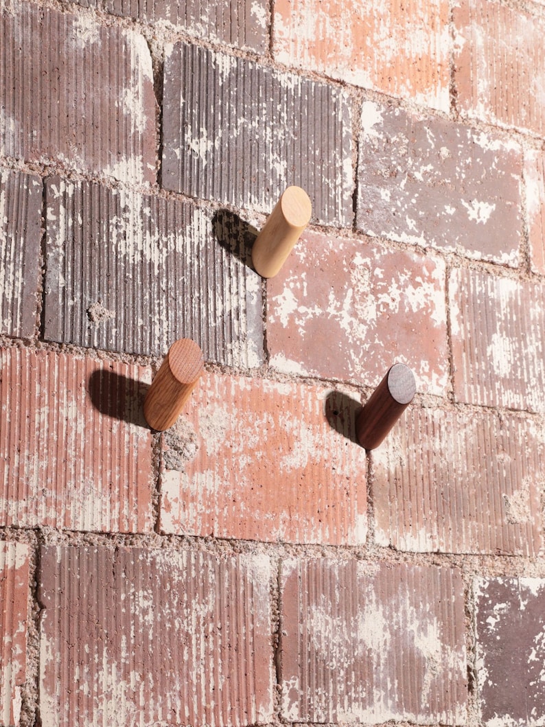 May include: Three wooden hooks mounted on a brick wall. The hooks are different colors: light brown, dark brown, and natural wood.