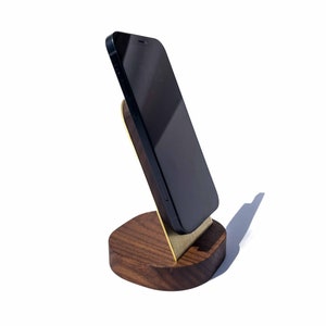 May include: A black smartphone is propped up in a wooden stand with a gold metal accent. The stand is made of dark wood and has a rounded base.