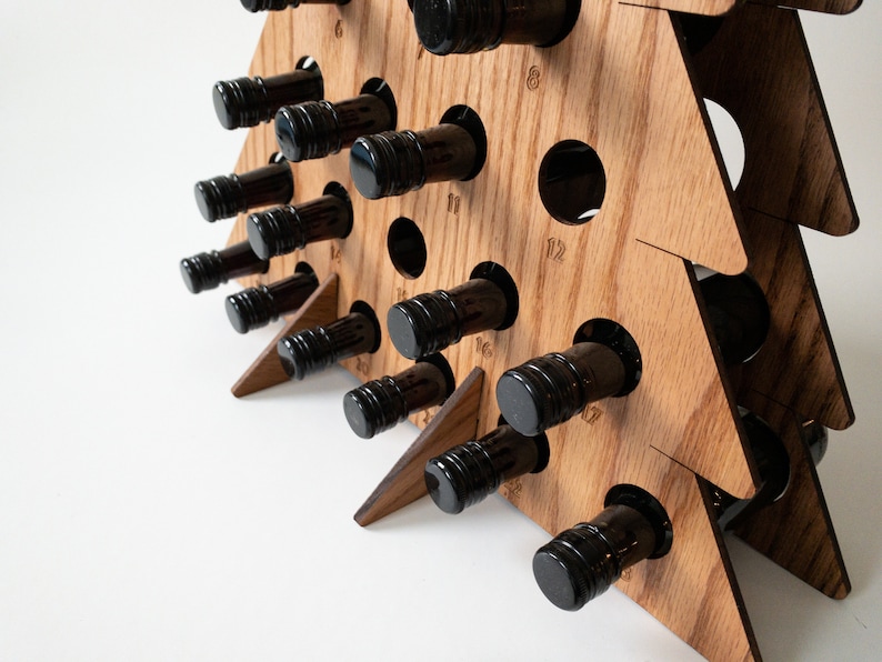 Puede incluir: Porta botellas de vino de madera con forma de &aacute;rbol de Navidad con 12 ranuras. El soporte est&aacute; hecho de madera marr&oacute;n claro y tiene un acabado de veta de madera natural. Las ranuras est&aacute;n numeradas del 1 al 12. El soporte est&aacute; sobre una superficie blanca.