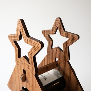 Puede incluir: Un estante de vino de madera con forma de casa con dos recortes en forma de estrella en el techo. El estante est&aacute; sosteniendo una botella de vino.