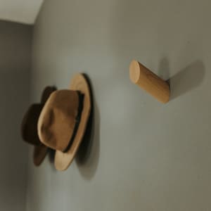 May include: Two brown felt hats hanging on a wall with a wooden hook. The hook is a cylinder shape and is mounted on a light gray wall.