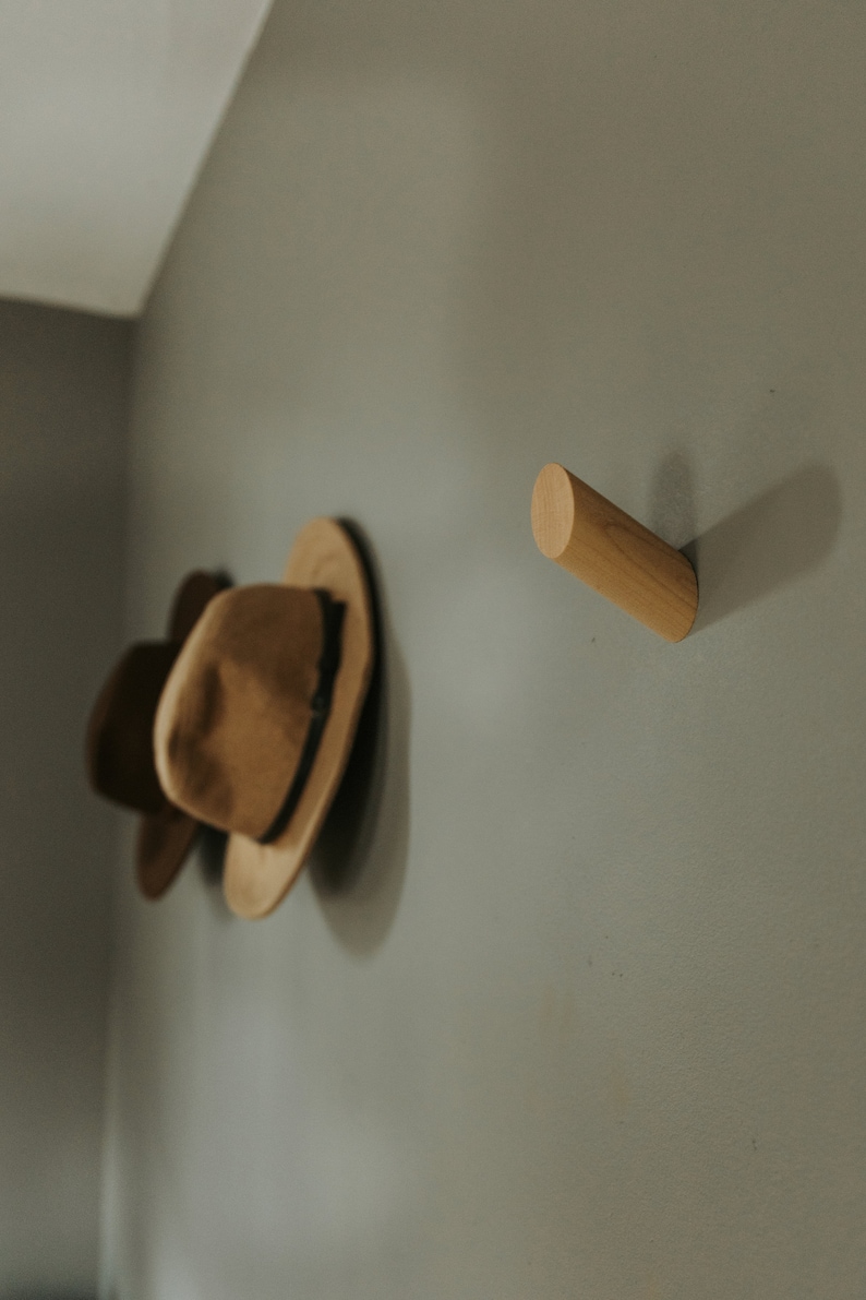 May include: Two brown felt hats hanging on a wall with a wooden hook. The hook is a cylinder shape and is mounted on a light gray wall.