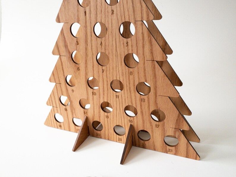 Puede incluir: Calendario de adviento de madera en forma de &aacute;rbol de Navidad con 23 c&iacute;rculos numerados. El &aacute;rbol est&aacute; hecho de madera marr&oacute;n claro y tiene una base triangular.