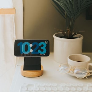 May include: A white charging stand with a black phone holder and a wooden base. A smartphone is on the stand displaying the time 10:23. A cup of coffee, a pair of glasses, and a plant are on a white table.